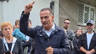 Un bărbat într-o jachetă albastră ridică degetul arătător în timp ce este înconjurat de mai multe persoane pe un fundal de clădire.