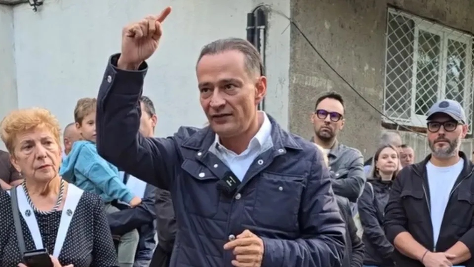 Un bărbat într-o jachetă albastră ridică degetul arătător în timp ce este înconjurat de mai multe persoane pe un fundal de clădire.