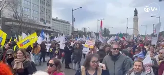 O mulțime de oameni cu diverse haine și steaguri colorate stă pe o stradă lângă clădiri înalte sub un cer înnorat.