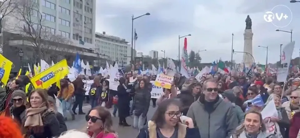 O mulțime de oameni cu diverse haine și steaguri colorate stă pe o stradă lângă clădiri înalte sub un cer înnorat.