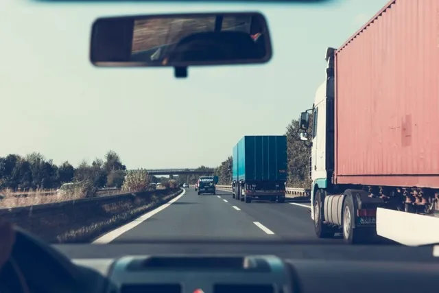 Privire de pe interiorul unei mașini spre un camion roșu și un camion albastru pe o autostradă cu vegetație în fundal.