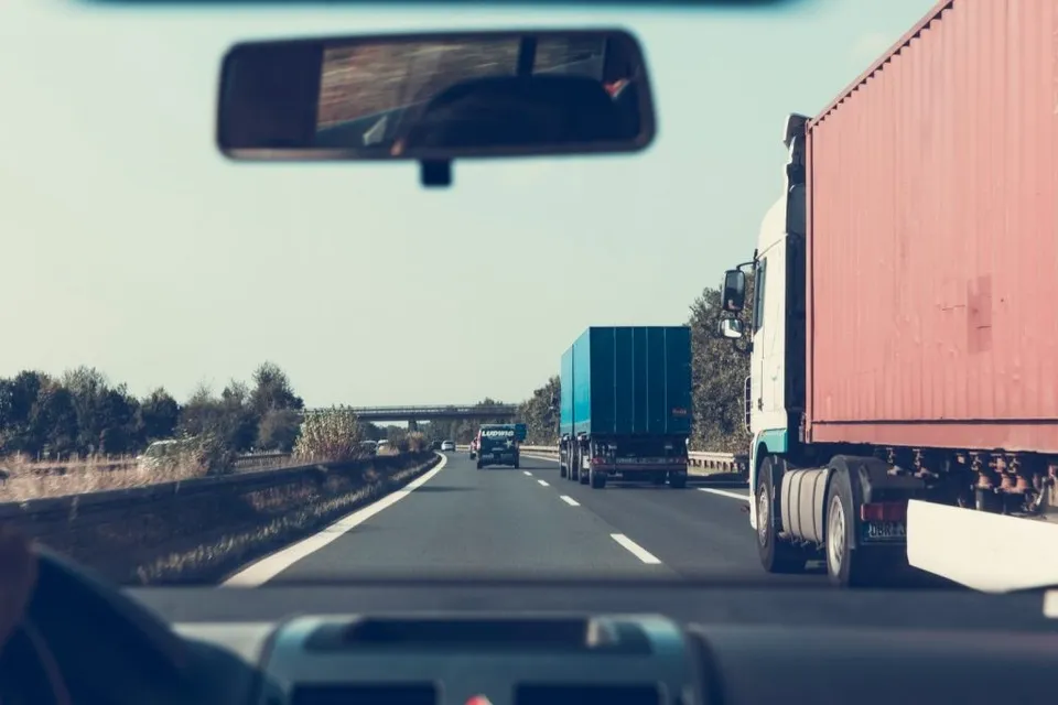 Privire de pe interiorul unei mașini spre un camion roșu și un camion albastru pe o autostradă cu vegetație în fundal.