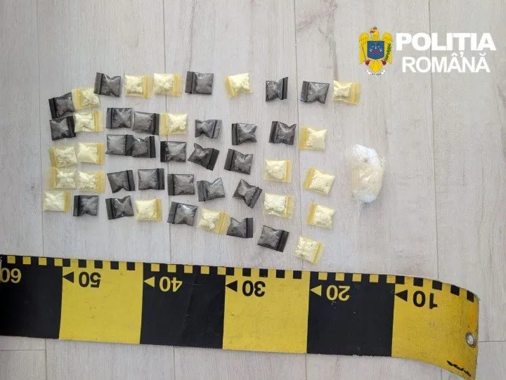 Multe pachete mici, unele în folie neagră și altele în folie transparentă, sunt așezate pe o suprafață plană lângă o bandă de măsură galbenă și neagră.