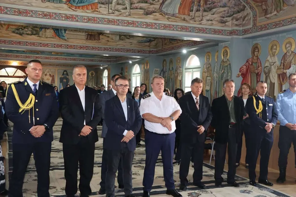 Un grup de bărbați stă în picioare în fața unui perete pictat cu figuri religioase într-o încăpere cu tavan înalt.