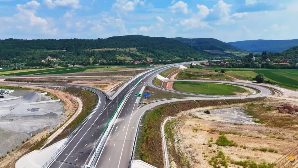 O autostradă cu mai multe benzi traversează un peisaj de dealuri verzi sub un cer cu nori albi.