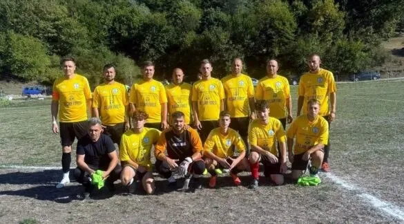 Un grup de oameni îmbrăcați în tricouri galbene și pantaloni scurți negri pozează pe un teren de fotbal cu vegetație în fundal.
