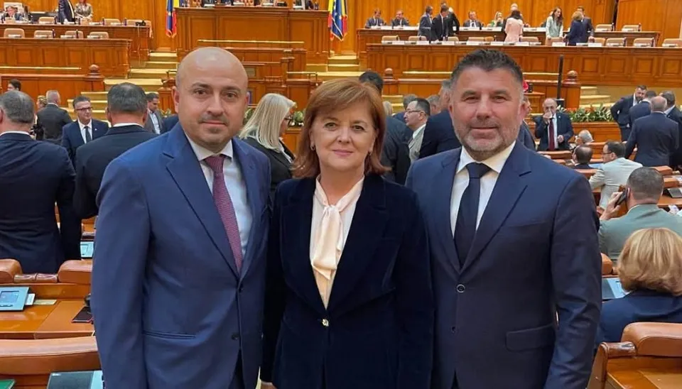 Trei persoane îmbrăcate în costume stau în picioare în fața unei săli cu scaune de lemn și mese de lucru.