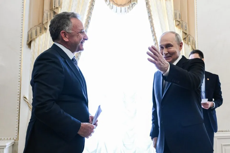Un bărbat într-un costum închis și un alt bărbat într-un costum închis stau în fața unei ferestre mari, unul ridicând mâna.