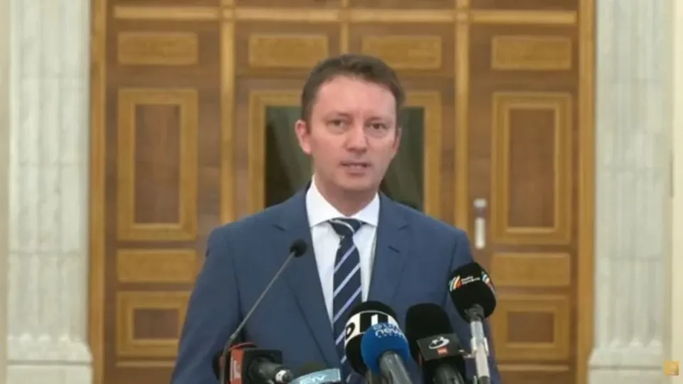 Un bărbat cu părul scurt, purtând sacou albastru, cămașă albă și cravată în dungi, stă în fața mai multor microfoane.
