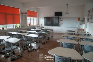 O sală de clasă cu mai multe birouri individuale cu picioare metalice gri și blaturi deschise dispuse pe un podea de lemn.