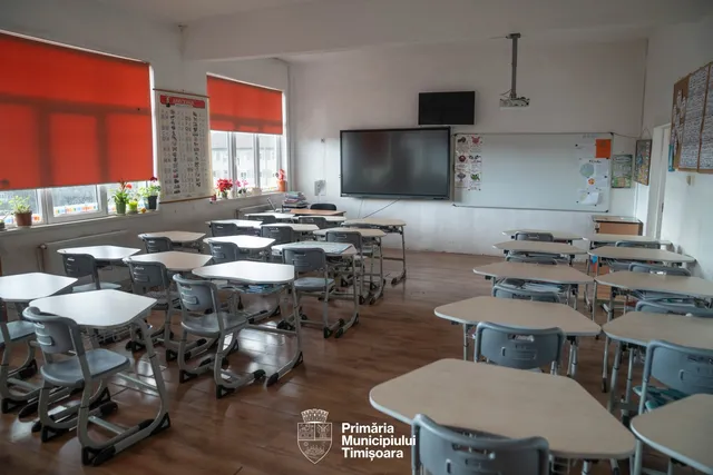 O sală de clasă cu mai multe birouri individuale cu picioare metalice gri și blaturi deschise dispuse pe un podea de lemn.