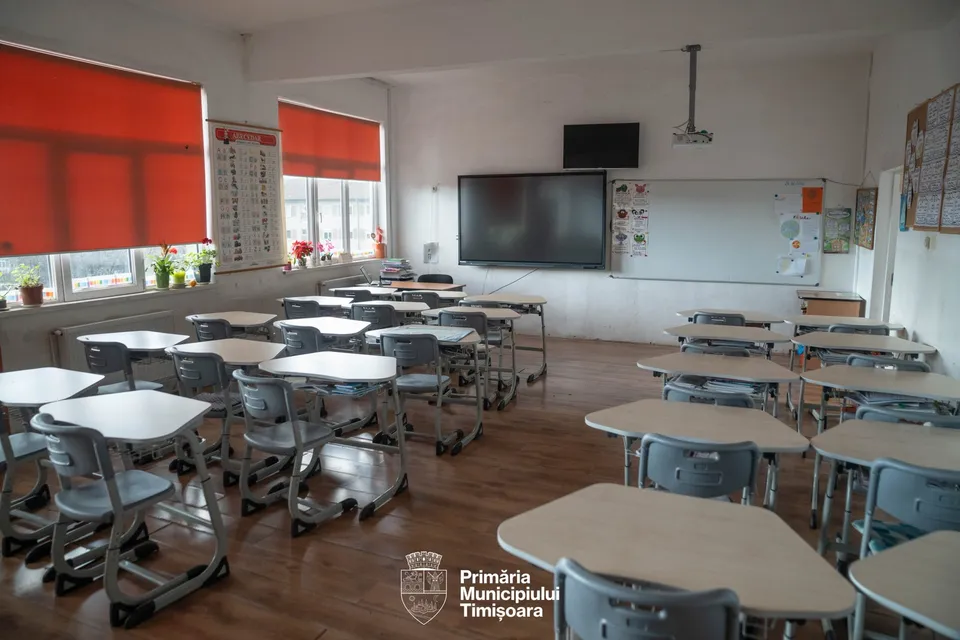 O sală de clasă cu mai multe birouri individuale cu picioare metalice gri și blaturi deschise dispuse pe un podea de lemn.