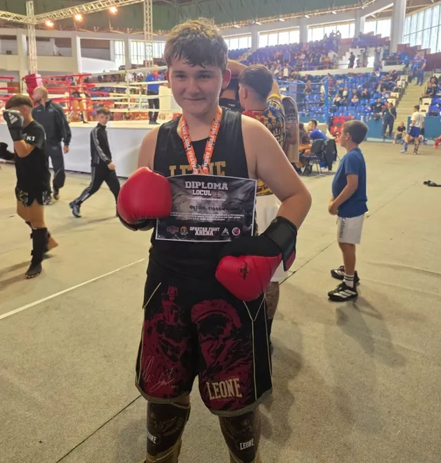 Un băiat poartă mănuși de kickboxing roșii, pantaloni de sport negri cu imprimeuri și un diploma în mână, stând într-o sală de sport cu tribune.