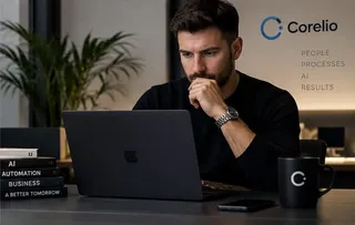 Un bărbat cu barbă și ceas de mână stă la o masă și privește un laptop deschis, având căni și cărți în apropiere.