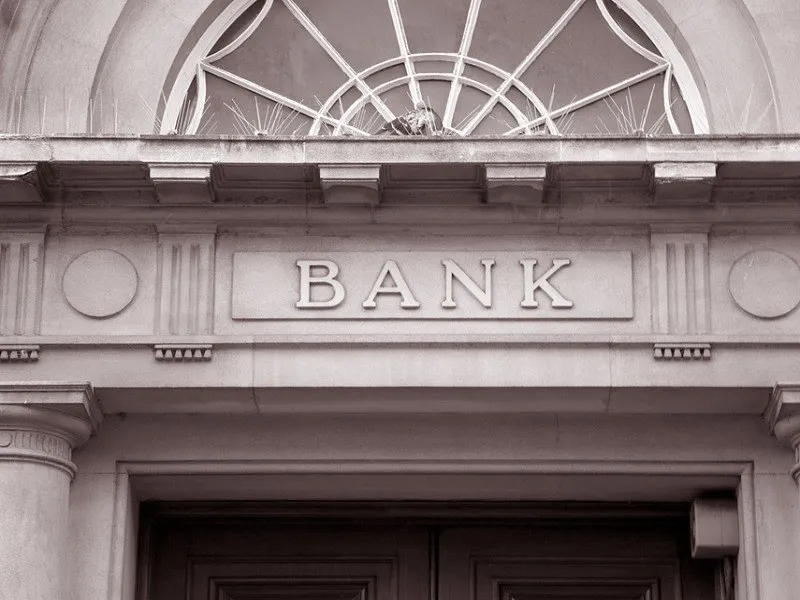 O fațadă de clădire cu inscripția BANK sculptată în piatră deasupra unei intrări cu coloane și un element decorativ circular deasupra.