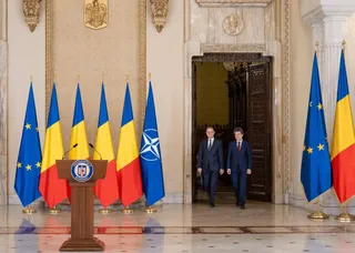 Două bărbați în costume întemeiate ies printr-o ușă deschisă, în timp ce în prim-plan sunt aliniate mai multe steaguri pe suporturi metalice.