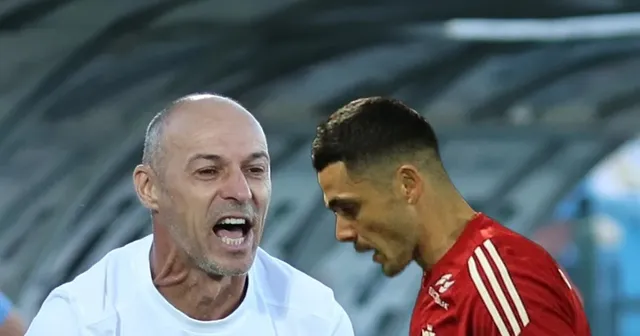 Un bărbat cu părul scurt și deschis la culoare poartă un tricou alb, în timp ce un alt bărbat cu părul scurt poartă un tricou roșu.