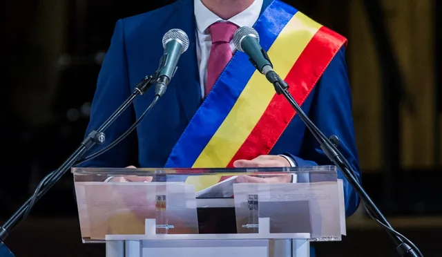 Un bărbat într-un costum albastru cu eșarfă tricoloră stă în spatele unui podium transparent cu două microfoane în față.