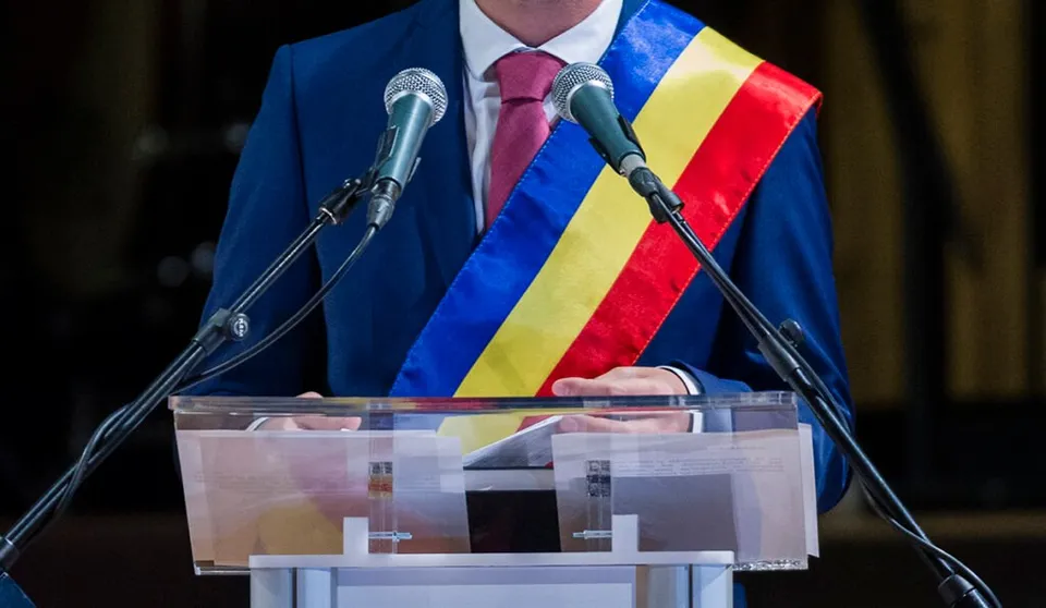 Un bărbat într-un costum albastru cu eșarfă tricoloră stă în spatele unui podium transparent cu două microfoane în față.