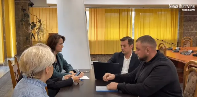 Patru persoane stau la o masă de lucru într-o încăpere cu jaluzele galbene, folosind un laptop și un telefon mobil.