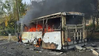 Un autobuz metalic este în flăcări și scoate fum dens pe un drum asfaltat lângă copaci.