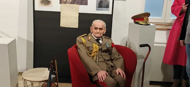 Un bătrân în uniformă militară cu epoleți și decoruri stă așezat într-un fotoliu roșu lângă un baston și un violoncel.