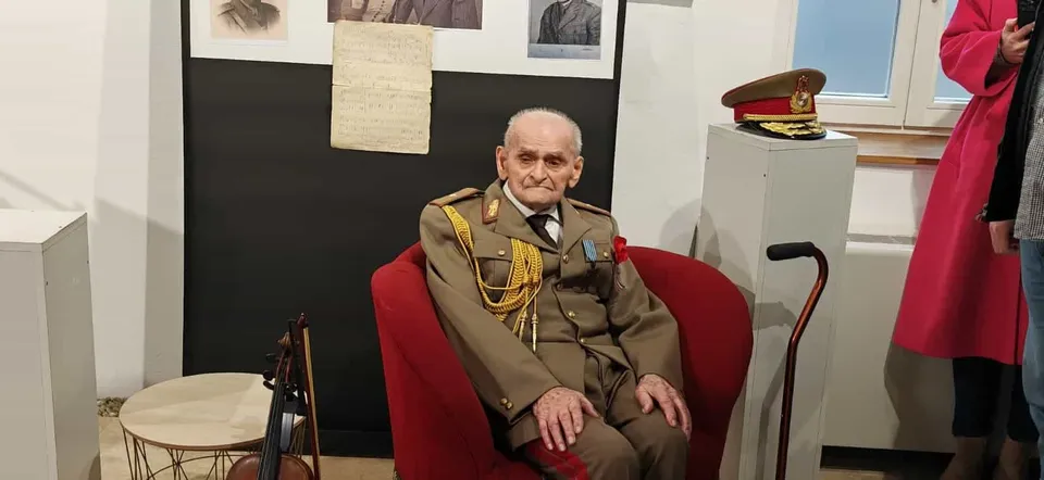 Un bătrân în uniformă militară cu epoleți și decoruri stă așezat într-un fotoliu roșu lângă un baston și un violoncel.