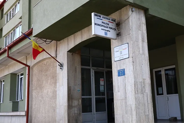 Intrarea unei clădiri este marcată de un panou alb cu text și un steag tricolor atașat pe un stâlp de perete.