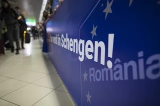 Un panou albastru cu textul scris în alb este situat lângă un hol de aeroport unde se văd persoane în fundal.