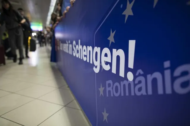 Un panou albastru cu textul scris în alb este situat lângă un hol de aeroport unde se văd persoane în fundal.