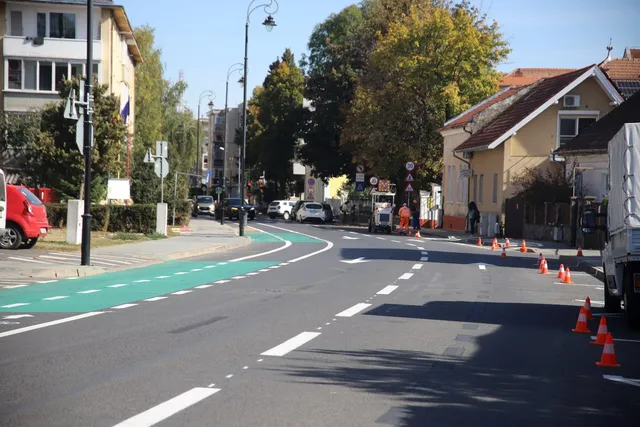 O bandă verde pentru bicicliști este marcată pe asfaltul unei străzi urbane lângă clădiri și copaci sub un cer senin.