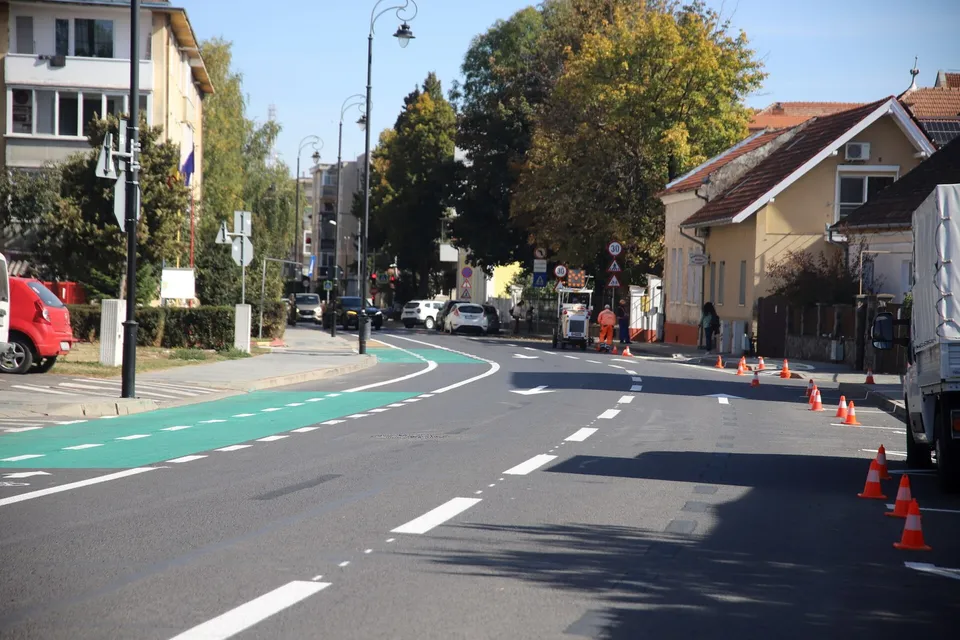 O bandă verde pentru bicicliști este marcată pe asfaltul unei străzi urbane lângă clădiri și copaci sub un cer senin.