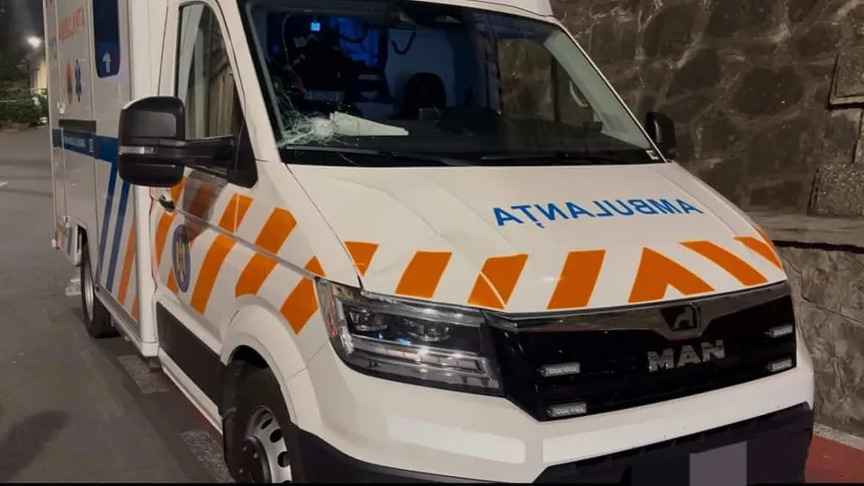 Un vehicul de tip ambulanță alb cu dungi portocalii și albastre este parcat pe lângă un perete de piatră.