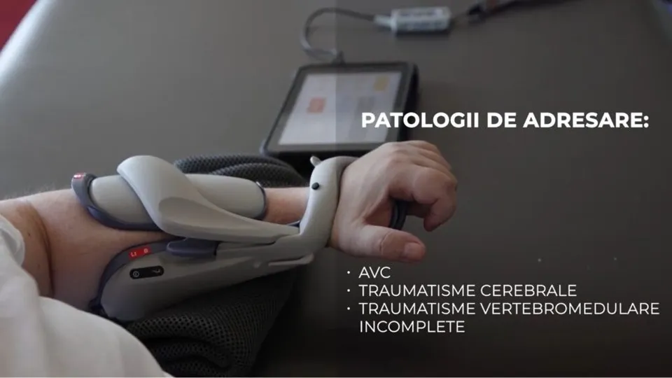 Un braț uman este poziționat pe o suprafață neagră, purtând un dispozitiv medical gri și alb cu cabluri conectat la o tabletă.
