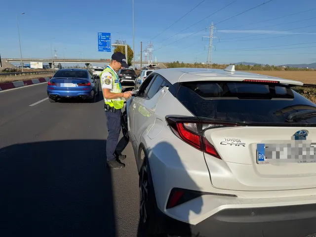 Un polițist în uniformă portivită scrie pe o foaie lângă o mașină albă pe marginea unui drum asfaltat sub un cer senin.