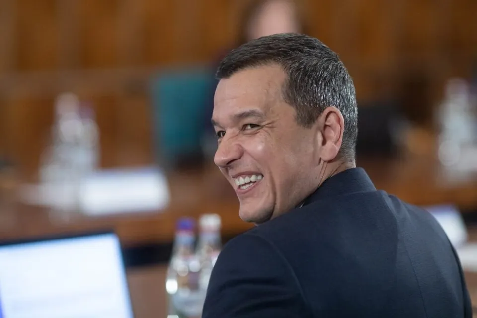 Un bărbat cu părul scurt și grij, purtând un sacou închis la culoare, zâmbește privind spre stânga într-un decor de birou cu obiecte desfocate.