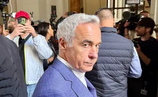 Un bărbat cu părul cărunt și sacou albastru este înconjurat de persoane care folosesc telefoane mobile și aparate foto într-un spațiu interior.