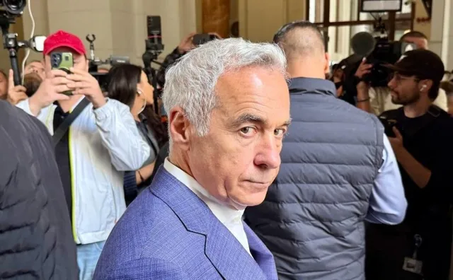 Un bărbat cu părul cărunt și costum albastru este înconjurat de persoane care dețin telefoane mobile și aparate foto într-un spațiu interior.