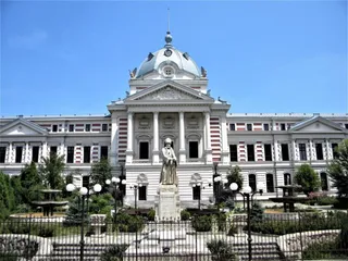 O clădire mare cu cupolă și coloane este situată în spatele unui monument sculptat și a unor grădini cu globuri de iluminat.