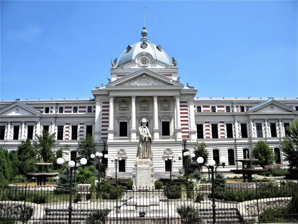 O clădire mare cu cupolă și coloane este situată în spatele unui monument sculptat și a unor grădini cu globuri de iluminat.