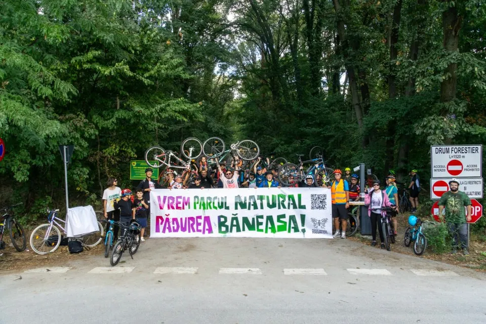 Un grup de oameni stă în fața unui banner alb cu text colorat, având numeroase biciclete ridicate deasupra capetelor în fața unei păduri.