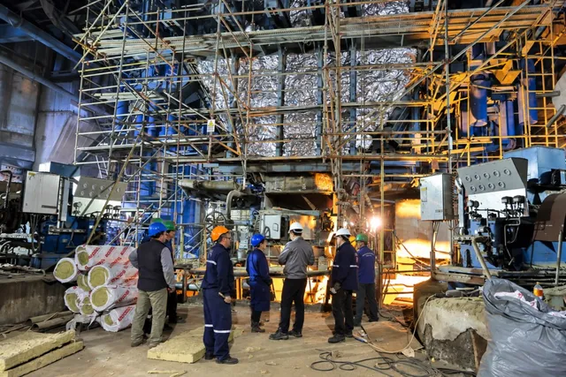 Mai mulți bărbați cu cască de protecție stau în fața unei structuri industriale mari cu schele și conducte metalice.