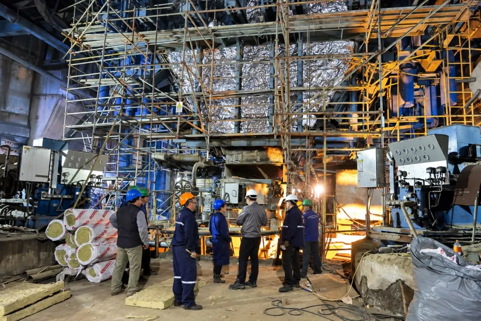 Mai mulți bărbați cu cască de protecție stau în fața unei structuri industriale mari cu schele și conducte metalice.