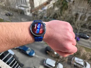 Un ceas inteligent cu brățară albastră este purtat pe un braț deasupra unui fundal urban cu mașini și clădiri.