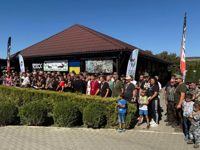 Un grup mare de oameni, inclusiv copii, stau în fața unei clădiri cu acoperiș maro, sub un cer senin. Unii poartă medalii.