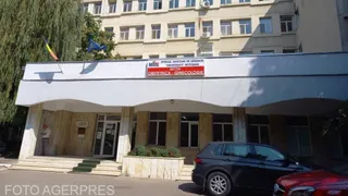 O clădire cu ferestre multiple prezintă o intrare acoperită sub care sunt parcate două mașini pe un parcare asfaltată.