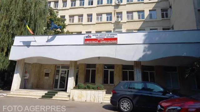 O clădire cu ferestre multiple prezintă o intrare acoperită sub care sunt parcate două mașini pe un parcare asfaltată.