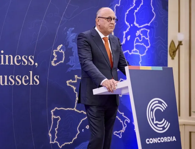 Un bărbat cu ochelari și costum închis la culoare stă în spatele unui podium alb cu logo-ul Concordia.