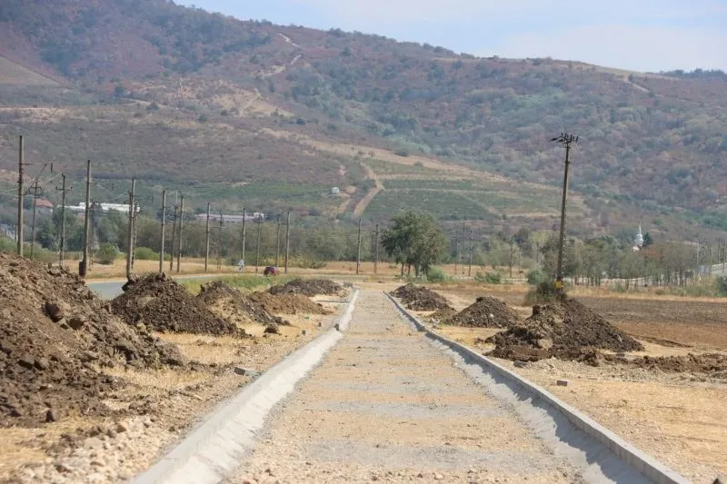 O cale pietruită cu borduri de beton se întinde spre un deal împădurit sub un cer senin.