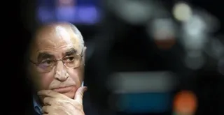 Un bărbat cu ochelari poartă un costum și își ține mâna sub bărbie, privind spre lateral într-un decor întunecat cu lumini difuze în fundal.